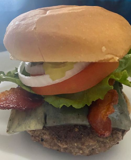 A close-up of a burger with lettuce, tomato, pickles, onions, bacon, and a soft bun on a white plate.