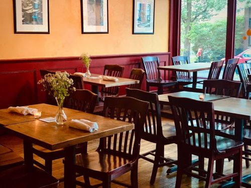 Cozy restaurant interior with wooden tables, chairs, and small flower arrangements on each table. Natural light from windows.