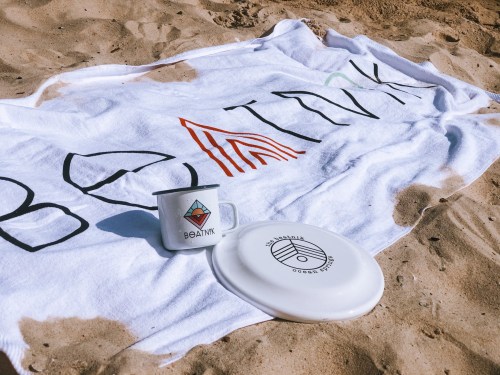 A beach scene featuring a white towel with "BEATINK," a mug, and a frisbee on sandy ground.