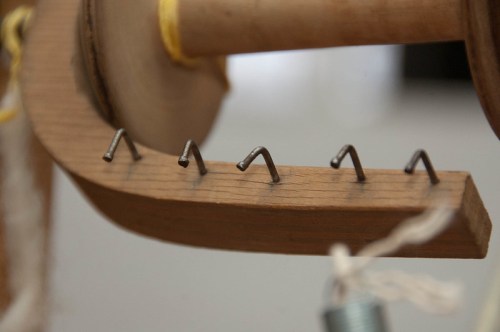 Close-up of a wooden spool with metal hooks arranged along its edge, with a thread partially visible.