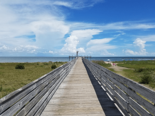 A wooden pier extends into the ocean under a bright blue sky with fluffy clouds and green grass on either side.