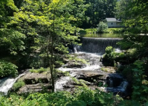 A serene waterfall cascades over rocks, surrounded by lush greenery and a small white building in the background.