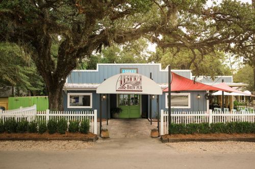 A charming brew pub with a green door, white picket fence, and large trees in a scenic outdoor setting.