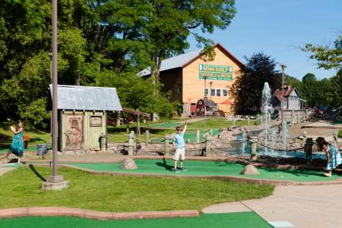 A mini-golf course with a fountain, colorful buildings, and players enjoying the game on a sunny day.