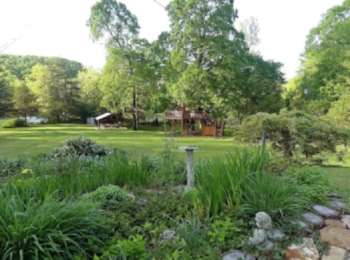 A lush green yard with a playset in the background, surrounded by trees and a small garden in the foreground.
