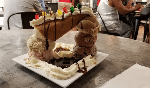 A large ice cream dessert featuring multiple scoops, whipped cream, a cone, chocolate drizzle, and colorful candy toppings.