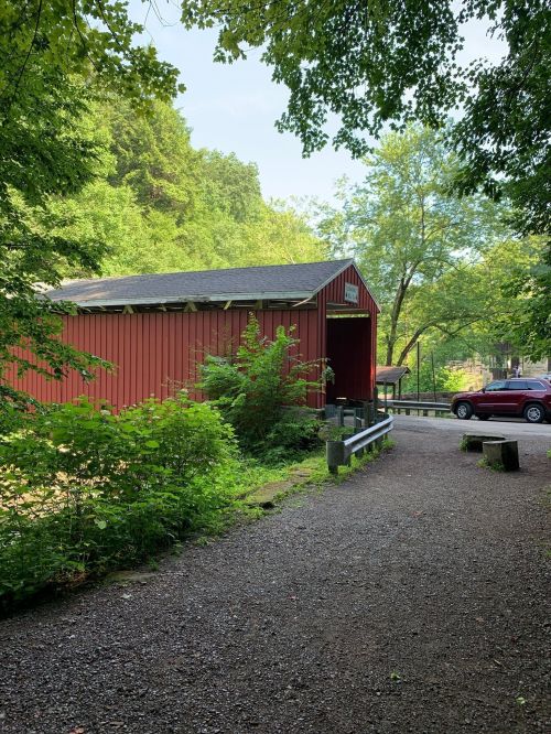 A red covered bridge surrounded by greenery, with a gravel path and a parked car nearby.