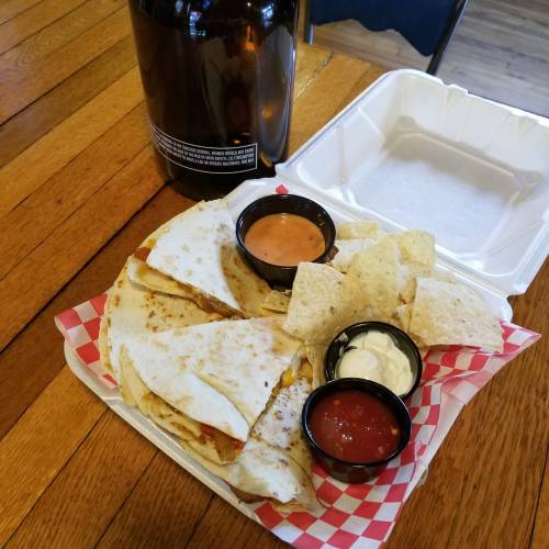 A takeout container with quesadillas, tortilla chips, and dipping sauces on a wooden table.