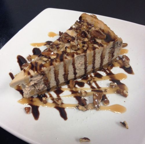 A slice of layered cheesecake drizzled with chocolate and caramel sauce, topped with chopped nuts on a white plate.