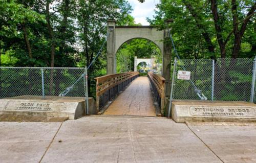 A wooden bridge leads through an archway, surrounded by trees, with signs indicating its history and park name.