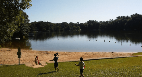 Two children play on a grassy area near a calm lake, with a sandy beach and trees in the background under a clear sky.