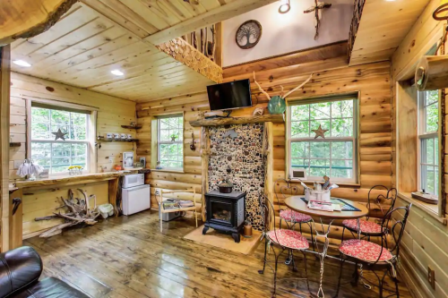 Cozy log cabin interior with wooden walls, a stone fireplace, dining table, and kitchen area, surrounded by large windows.