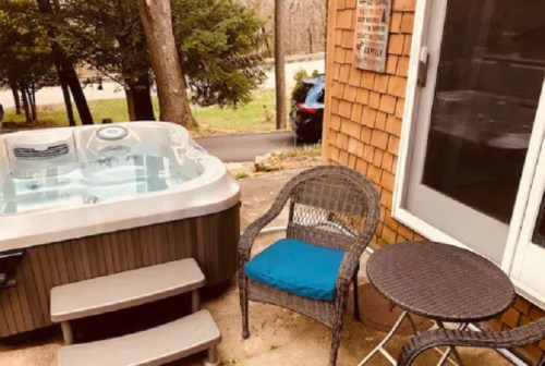 A hot tub on a patio next to a small table and chair, surrounded by trees and a view of a road.