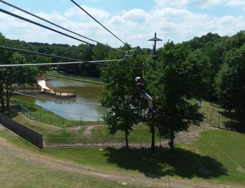 A person zip-lining over a pond surrounded by trees, with a wooden platform in the background under a blue sky.