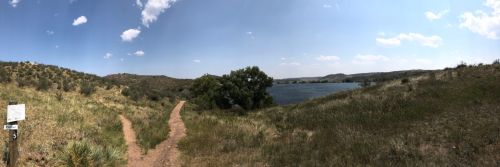 A panoramic view of a grassy landscape with a winding path leading to a lake surrounded by hills and blue sky.