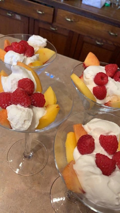 Four dessert cups filled with peach slices, whipped cream, and fresh raspberries on a countertop.