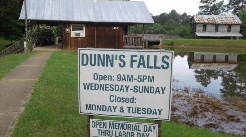 Sign for Dunn's Falls with hours of operation, a wooden building, and a pond in the background. Closed Monday and Tuesday.