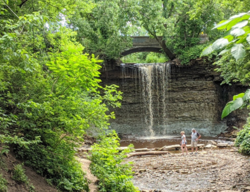 A serene waterfall cascades over rocky cliffs, surrounded by lush greenery, with two people exploring the area below.