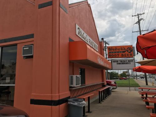 Exterior of a pink restaurant with a sign for root beer and burgers, featuring outdoor seating and a cloudy sky.