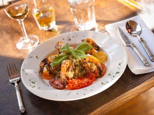 A plate of grilled shrimp on rice, garnished with basil and served with sliced tomatoes, alongside glasses of drink.