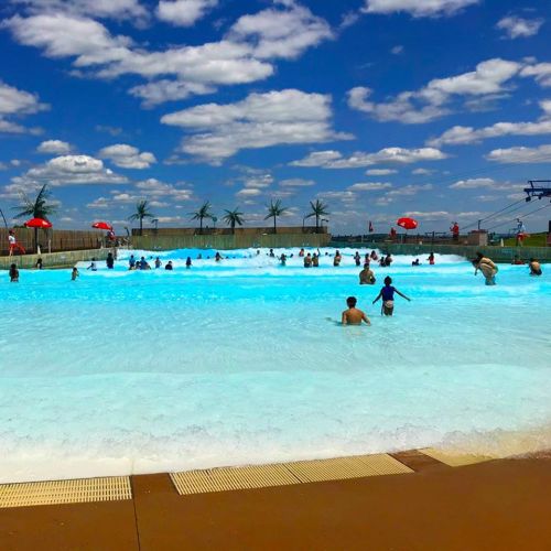 A vibrant wave pool filled with people enjoying the water under a blue sky with fluffy clouds and palm trees.