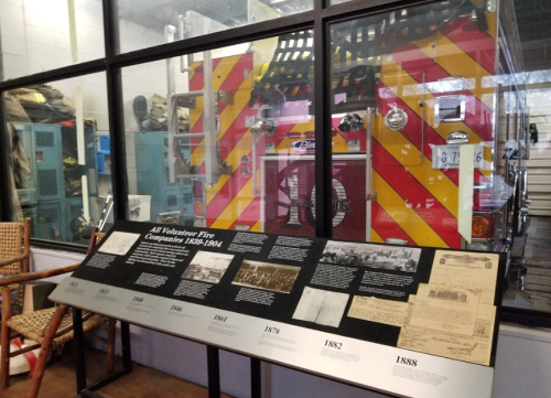 A fire truck behind glass, with a display of historical fire company documents and photos in the foreground.