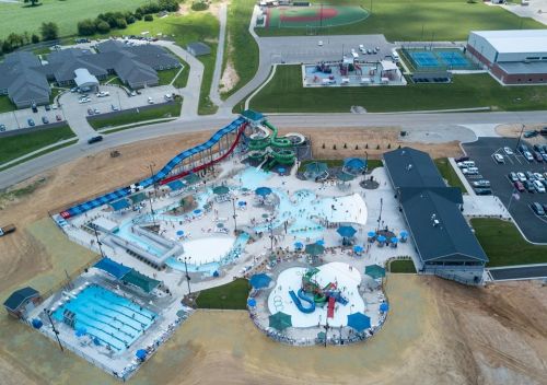 Aerial view of a large water park with pools, slides, and recreational areas, surrounded by green fields and buildings.