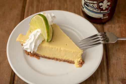 A slice of lime cheesecake topped with whipped cream and a lime wedge on a white plate, with a drink in the background.