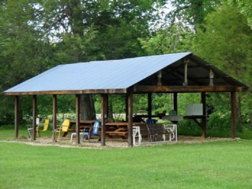 A wooden pavilion with a blue roof, surrounded by green grass and chairs, set in a wooded area.