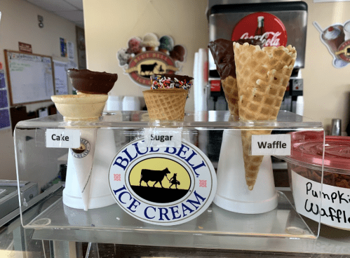 Display of ice cream cones labeled "Cake," "Sugar," and "Waffle" in a shop, with a Blue Bell Ice Cream logo.
