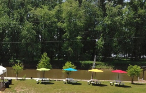 Colorful umbrellas shade picnic tables by a calm river, surrounded by lush green trees on a sunny day.