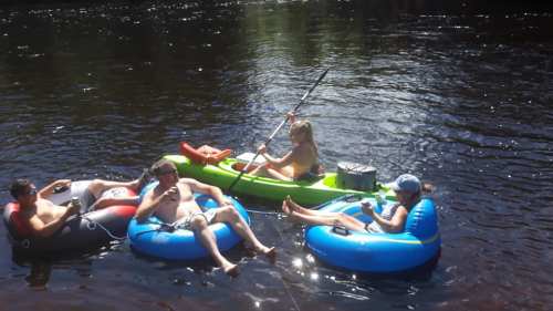 Four people relax on inflatable rafts in a river, with one person kayaking nearby on a sunny day.
