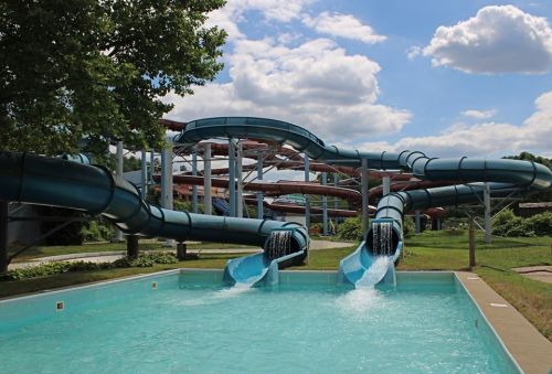 Two water slides descend into a clear pool, surrounded by green grass and trees, under a blue sky with fluffy clouds.