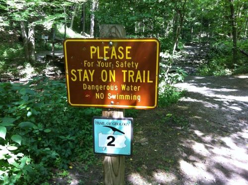 A brown sign in a wooded area warns to stay on the trail due to dangerous water and prohibits swimming.