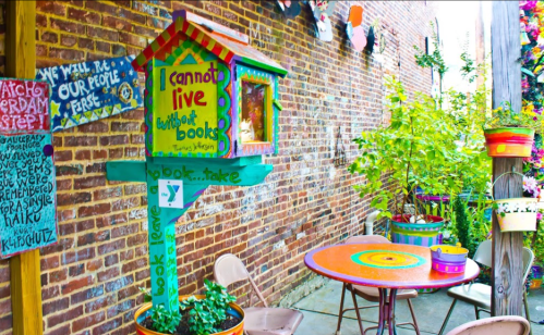 Colorful book exchange box in a vibrant outdoor space with a round table and plants against a brick wall.