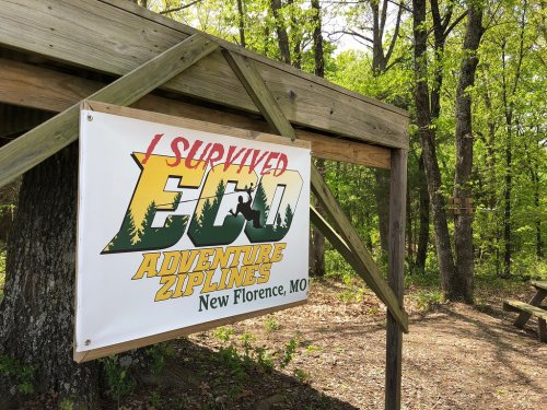A banner reading "I Survived Eco Adventure Zip Lines" hangs under a wooden structure in a wooded area.