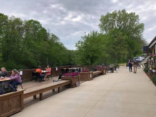 Outdoor dining area with wooden tables, flowers, and trees, featuring people enjoying meals and a cloudy sky.