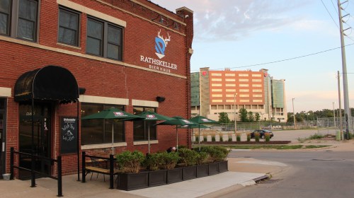 Exterior of RathsKeller Bier Haus with outdoor seating and green umbrellas, set against a cityscape backdrop.