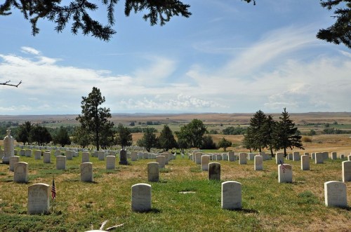 A serene cemetery with white gravestones, surrounded by green grass and trees, under a blue sky with scattered clouds.