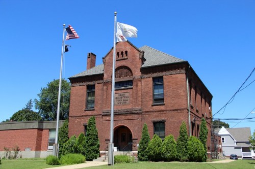 Historic brick building with flags in front, surrounded by greenery and a clear blue sky.