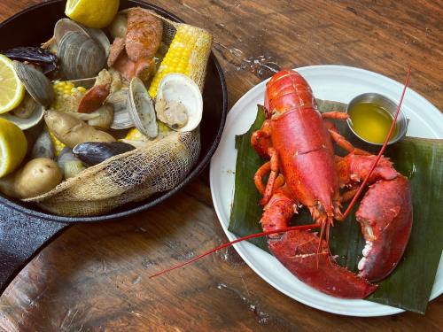 A cast iron skillet filled with seafood, corn, and potatoes, alongside a whole lobster on a plate with dipping sauce.
