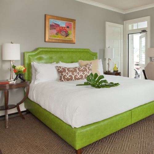 Bright bedroom featuring a green upholstered bed, white linens, decorative pillows, and a small table with a plant.
