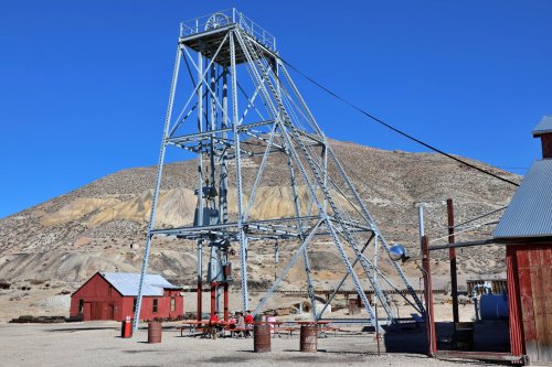 A tall metal mining structure stands beside red wooden buildings against a clear blue sky and mountainous backdrop.