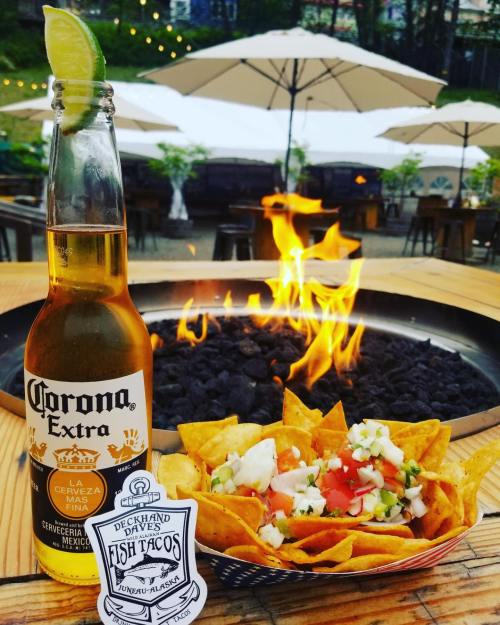A bottle of Corona beer next to a bowl of nachos with toppings, set against a fire pit backdrop.