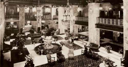 A grand hotel lobby featuring marble columns, chandeliers, a fountain, and elegant furnishings, adorned with flowers.