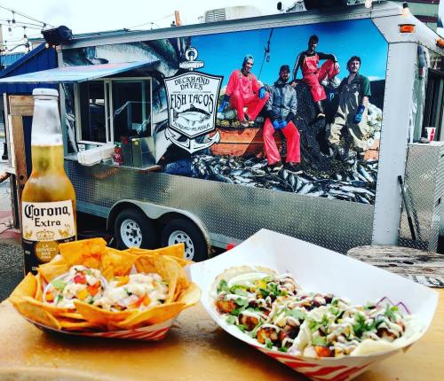 A food truck serving fish tacos, with tacos and nachos in the foreground and a scenic mural of fishermen in the background.
