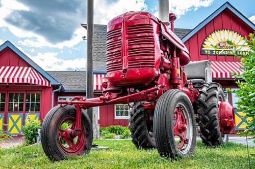 A bright red vintage tractor parked in front of a colorful barn with striped awnings and green landscaping.