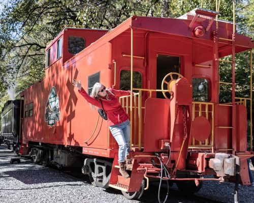 A woman in a red jacket playfully poses on the steps of a bright red train caboose surrounded by trees.