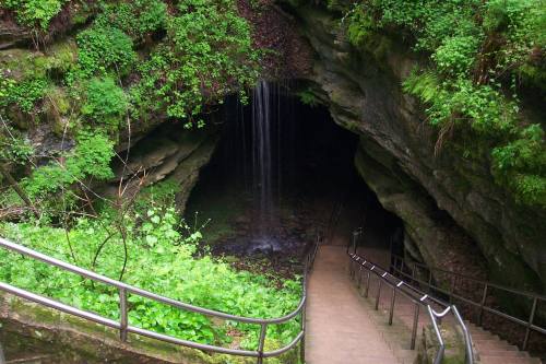 A lush green cave entrance with a small waterfall and stone steps leading down into the darkness.