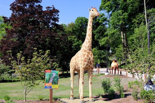 A tall giraffe statue stands in a park, surrounded by greenery and another animal statue in the background.